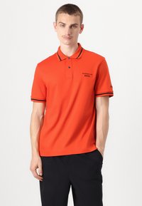 Raudona polo marškinėliai su trimis mygtukais, juodais akcentais ant apykaklės ir rankovių, bei logotipu ant krūtinės. Pagaminti iš glotnaus audinio.