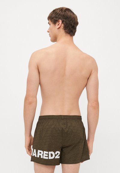 Junger Mann, der mit dem Rücken zur Kamera steht, trägt dunkelbraune Badeshorts mit wiederholtem Markentext und großem weißen "DSQUARED2"-Logo am linken Bein.