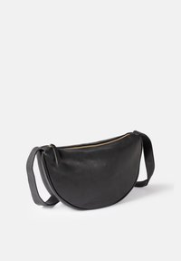 Black half-moon leather shoulder bag with gold zipper and adjustable strap, resting on a white surface.