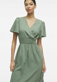 Zielona sukienka kopertowa z krótkimi bufkami i marszczoną talią. Gładki materiał, zwiewna spódnica i głęboki dekolt w kształcie litery V.