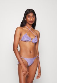 Conjunto de biquíni roxo com um top triangular texturizado com bordas franzidas e um detalhe em tom de ouro, combinado com calções de cintura baixa ajustáveis.