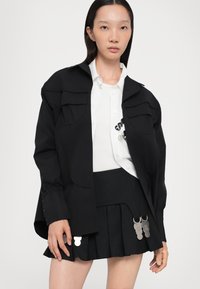 Chaqueta negra oversized con dos bolsillos en el pecho, superpuesta a una camisa blanca. La falda negra presenta pliegues y detalles en metal plateado.