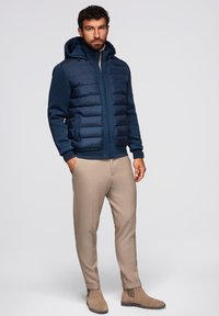 Giacca a vento blu navy con cappuccio, caratterizzata da una texture trapuntata e tasche laterali, abbinata a pantaloni beige su misura e stivaletti marroni alla caviglia.