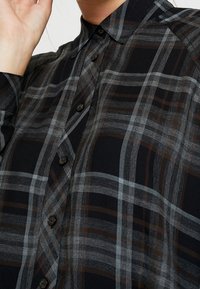 Schwarz-graues kariertes Hemd mit Knopfleiste, langen Ärmeln und Kragen. Besitzt braune und hellblaue Linien im Muster.