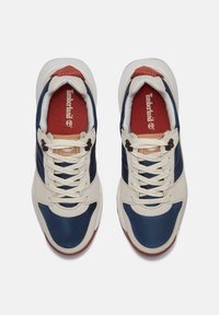 Vita och marinblå sneakers med en blandad textilovandel, platta snören, gummisula och röd innersula med "Timberland"-logotyp.