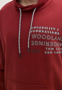 Czerwona bluza z kapturem z szarymi sznurkami. Na przodzie znajduje się tekst drukowany w różnych czcionkach i rozmiarach. Tekst zawiera słowa "TRWAŁOŚĆ" i "1962".