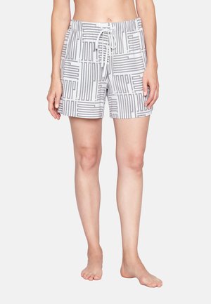 Žena v bílých šortkách se šedým geometrickým pruhem a stahovací šňůrkou v pase, stojící bosá na jednobarevném pozadí.