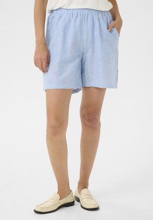Shorts tissés bleu clair avec une taille élastique, des poches latérales et une coupe décontractée. Portés avec des mocassins crème.