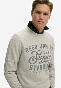 Joven con cabello corto que viste un suéter gris claro con texto impreso y una camisa de cuadros oscura debajo, sobre un fondo blanco liso.