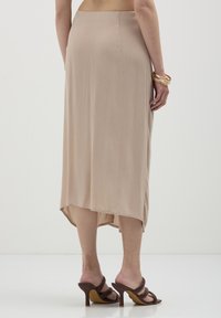 Jupe beige, longueur genou, fabriquée en tissu léger, avec un motif texturé subtil et un ourlet arrière légèrement plus long. Associée à des sandales à talons hauts marron.