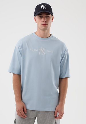 Mężczyzna ubrany w jasnoniebieską koszulkę New York Yankees i czarną czapkę Yankees, stojący na tle jednolitego białego tła.