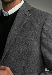 Szary żakiet w jodełkę z notched lapel, pojedynczą kieszenią na piersi i widocznym szwem, noszony na czarnym swetrze i białej koszuli.
