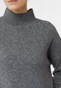 Nahaufnahme einer Person, die einen grauen gerippten Rollkragenpullover mit einer weichen strukturierten Masche und teilweise langen Ärmeln trägt.