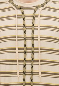 Μπεζ φούτερ με λεπτές οριζόντιες ρίγες σε καφέ και κίτρινο, διαθέτει δύο μπεζ κορδόνια και δύο πιο χοντρά, μοτίβο σε μπεζ και μαύρο κορδόνια.