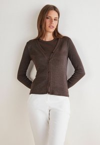 Frau in braunem, geknöpftem Cardigan über passendem Oberteil und weißen Hosen, steht mit den Händen hinter dem Rücken vor einer schlichten Wand.