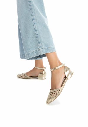 Zapatos planos metálicos dorados con diseño cortado, puntera afilada y tira alrededor del tobillo. Presentan un pequeño tacón en bloque transparente. Combinados con pantalones de mezclilla anchos.