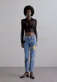 Črna prosojna srajca z dolgimi rokavi in gumbi, oprijeta v pasu, v kombinaciji s svetlo modrimi jeans hlačami, okrašenimi z rumenimi in črnimi grafičnimi motivi.