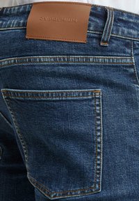 Tummansiniset farkut, joissa on saumaton pinta. Vyötäröllä on ruskea nahkapläntti, jossa on kohokuvioitu teksti, sekä näkyvää ompelua.