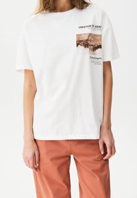 T-shirt en coton blanc avec un design imprimé représentant "La Création d'Adam" de Michel-Ange sur le devant, accompagné d'un texte décrivant l'œuvre.