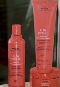 Aveda Nutri Plenish schampo och balsam i röda flaskor, med en jämn textur och vattendroppar på ytan för återfuktning.