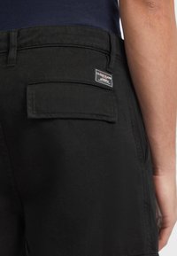 Crne traper kratke hlače imaju stražnji džep s kvadratnom oznakom Guess Jeans. Tkanina je glatka s laganim sjajem.