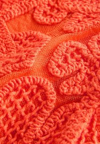 Close-up af lyst orange hæklet stof med hævede, hvirvlende mønstre på en fin maskebaggrund.