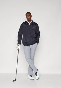 Marineblauwe pulloverjack met een halve rits, lichtgrijze slim-fit broek en witte golfschoenen met blauwe accenten; golfclub in de hand.
