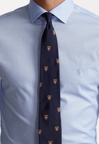 Tmavě modrá kravata s opakujícím se vzorem štítů ve zlaté, červené a bílé barvě, nošená s světle modrou texturovanou košilí s jemným logem.