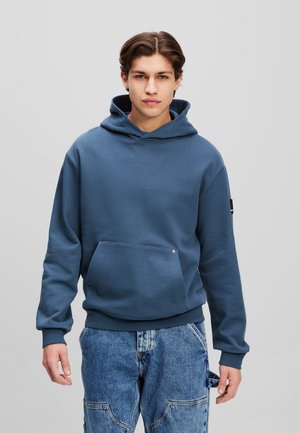 Blå sweatshirt med huva, tillverkad av mjukt tyg. Har en framficka i känguru-stil och ribbade ärmmuddar. Kombinationen med löst, urblekta denimbyxor.