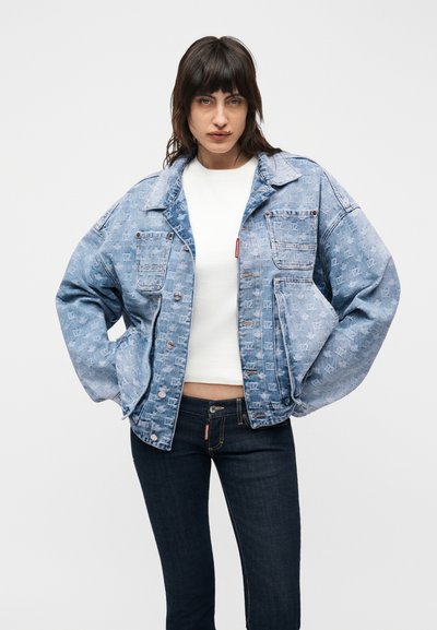 Femme aux cheveux foncés portant une veste en jean oversize bleu clair sur un crop top blanc et un jean bleu foncé, debout les mains dans les poches.