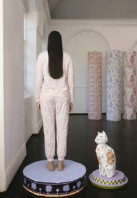 Een persoon met lang haar, gekleed in een pyjama met een patroon, staat op een rond platform naast een geschilderde katten sculptuur in een galerie met versierde zuilen.