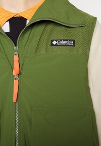 Veste verte Columbia Sportswear avec curseurs de fermeture éclair orange portée sur un haut noir et blanc avec un col orange.