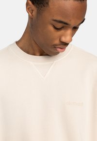 Sudadera beige claro con cuello redondo acanalado y un sutil detalle de diseño en forma de V en el escote. Cuenta con un pequeño logo bordado en el pecho.