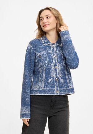 Frau mit blondem Haar, die eine blau-weiß gemusterte Jacke mit Knöpfen und schwarze Hose trägt, schaut mit einem leichten Lächeln nach rechts.