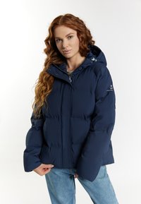 Jachetă puffer albastră marin cu glugă și închidere cu fermoar. Are un design cusut, manșete elastice și un logo pe piept. Pur și simplu cu blugi albaștri deschisi.