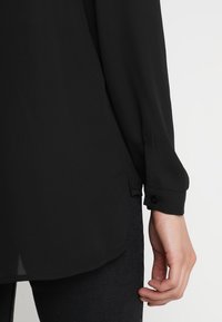 Maglietta nera a maniche lunghe con una texture liscia, orlo arrotondato e polsini con bottoni, abbinata a jeans scuri, che mostrano un design semplice e classico.