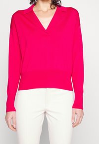 Pull-over en maille fuchsia avec col en V et col, manches longues avec poignets côtelés, associé à un pantalon blanc, avec une coupe décontractée.