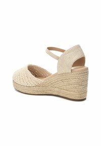 Sandale compensée en espadrille beige avec texture tissée, bout ouvert, bride cheville et semelle en jute détaillée. Présente une doublure intérieure lisse.
