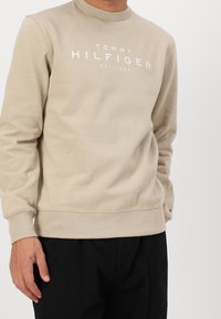 Sudadera beige con cuello redondo, mangas largas y logo blanco de "TOMMY HILFIGER". Tejido suave y puños y dobladillo acanalados. Pantalones negros visibles en la parte inferior.