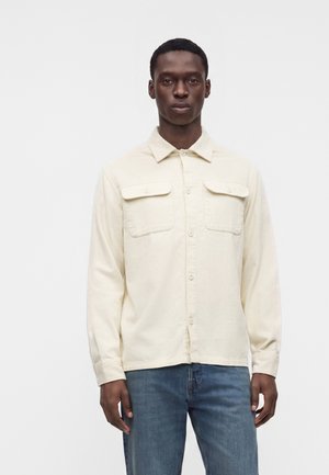 Homme debout, portant une chemise crème boutonnée avec poches sur le devant et un jean bleu, de face sur un fond blanc uni.