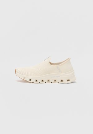 Zapatillas slip-on beige con un upper texturizado, suela acolchada y un diseño de suela acentuado que presenta recortes circulares. Branding minimalista visible.