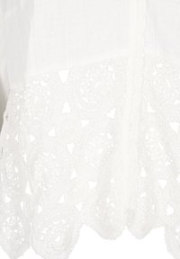 Vit topp med intrikata virkat spetsdetaljer och cirkulära mönster längs fållen. Lättviktsmaterial med en texturerad yta.