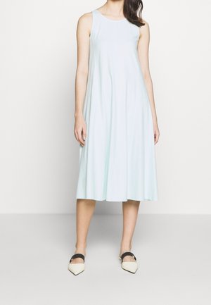 Vrouw draagt een mouwloze lichtblauwe midi-jurk met een ronde halslijn en witte platte schoenen met een spitse neus en zwarte bandjes.