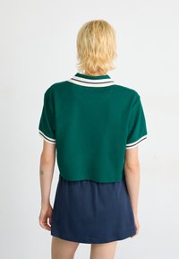 Grønn kortermet polohett med hvite og marineblå striper, sammen med et mørkeblått skjørt. Glatt stoff og avslappet passform.
