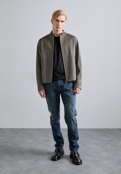 Grå teksturert jakke med høy krage, parret med mørke jeans og skinnende svarte sko, mot en lys grå bakgrunn.