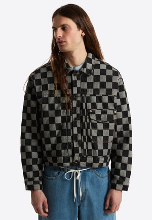 Hombre con pelo largo que lleva una chaqueta de botones a cuadros en negro y gris y vaqueros azules con cordón blanco.