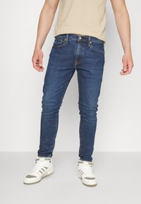 Modré úzké džínové kalhoty z denimu s pětikapsovým designem, jemnými švy a středně vysokým pasem, kombinované s bílými vysokými teniskami.