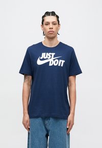 Μπλουζάκι από βαμβάκι σε ναυτικό μπλε χρώμα με κοντά μανίκια, διακοσμημένο με μεγάλο λευκό λογότυπο "Just Do It" της Nike στο στήθος. Άνετη γραμμή.