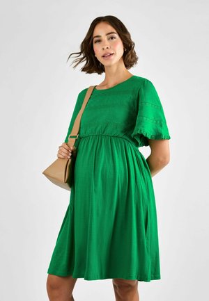 Abito verde a maniche corte con tessuto texturizzato, corpino aderente e gonna svasata; abbinato a una borsa a spalla color sabbia.