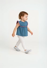 Bambino con capelli ricci che cammina, indossando una maglietta senza maniche blu con bordi rosa, leggings bianchi e sneaker bianchi su uno sfondo neutro.
